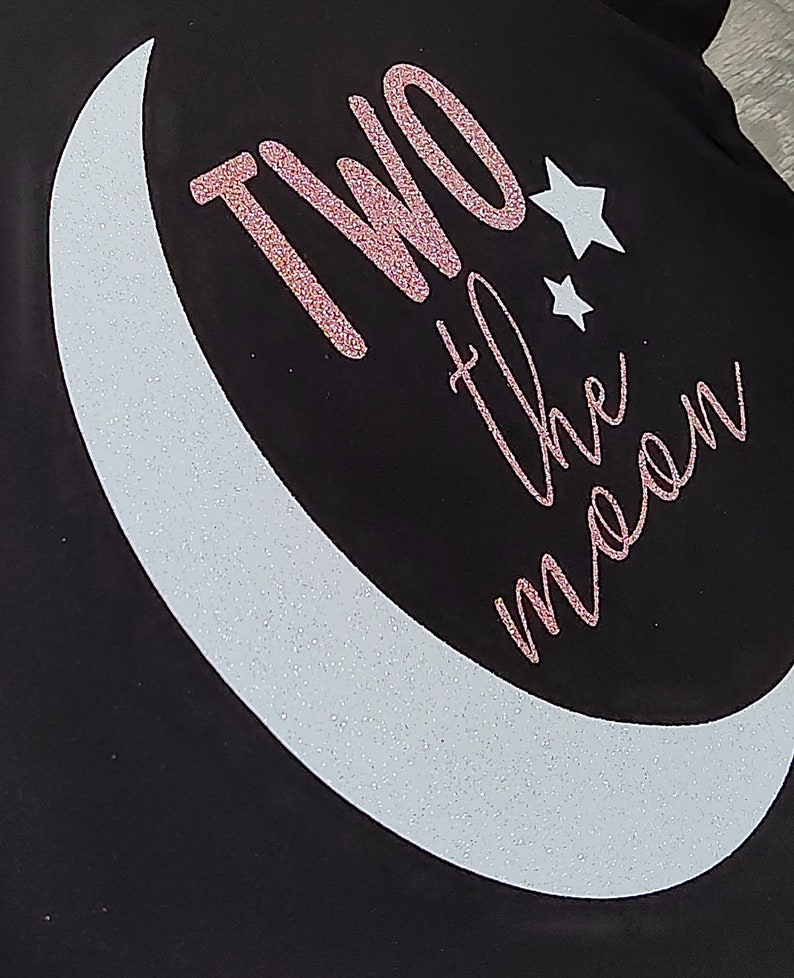 May include: Black fabric with a white crescent moon and two stars. The phrase "TWO the moon" is written in pink glitter script. The design is likely for apparel or accessories.