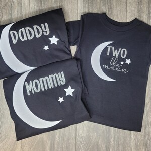 May include: Black t-shirts with white crescent moon and star designs. One shirt says "Daddy", another says "Mommy", and the third says "Two the moon". The shirts are folded and arranged on a wooden surface.
