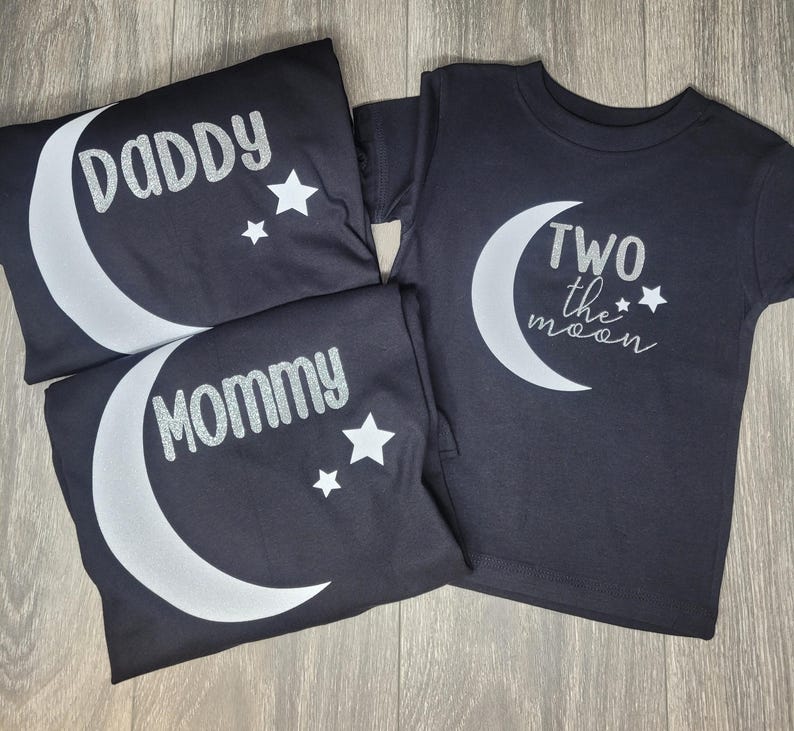 May include: Black t-shirts with white crescent moon and star designs. One shirt says "Daddy", another says "Mommy", and the third says "Two the moon". The shirts are folded and arranged on a wooden surface.