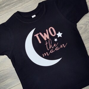 May include: Black toddler t-shirt with a white crescent moon and the phrase "Two the moon" in rose gold glitter and cursive font. Two small white stars are also included in the design.