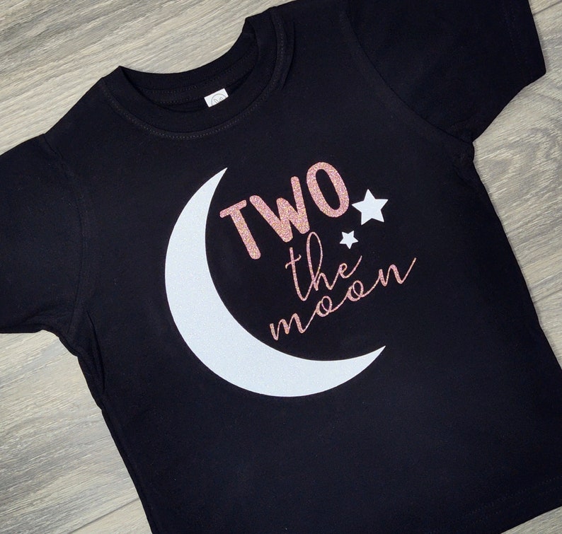 May include: Black toddler t-shirt with a white crescent moon and the phrase "Two the moon" in rose gold glitter and cursive font. Two small white stars are also included in the design.