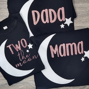 May include: Black t-shirts with white crescent moon designs and rose gold glitter text. One shirt says "DADA", another says "MAMA", and a third says "Two the moon". Each shirt has white stars.