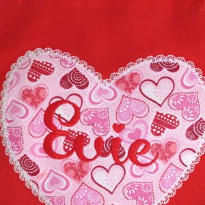May include: Red fabric tote bag with a white heart-shaped patch. The patch is covered in pink and red hearts and the word "Evie" is embroidered in red on the patch.