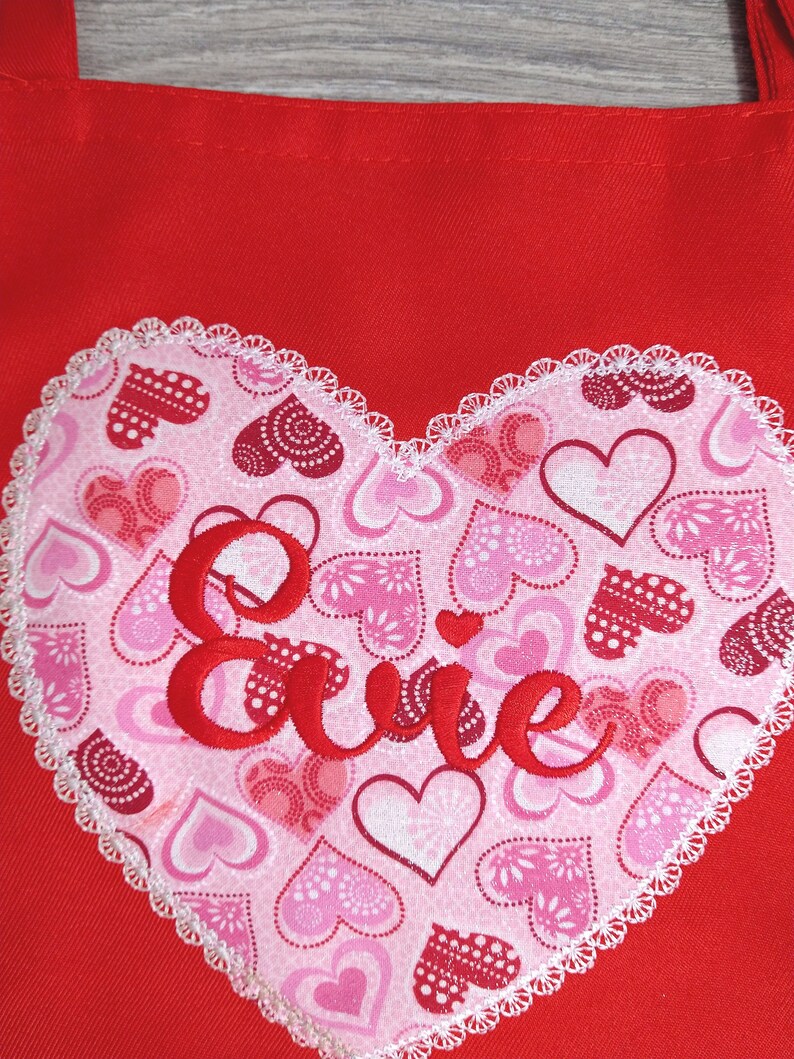 May include: Red fabric tote bag with a white heart-shaped patch. The patch is covered in pink and red hearts and the word "Evie" is embroidered in red on the patch.