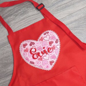 May include: Red apron with a large heart-shaped patch featuring a white lace trim and a pink and red heart pattern. The word "Love" is embroidered in red on the heart.
