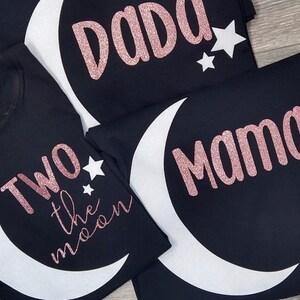 May include: Black t-shirts with white crescent moon and star designs. One shirt says "Two the moon" in pink glitter, another says "Dada", and a third says "Mama".