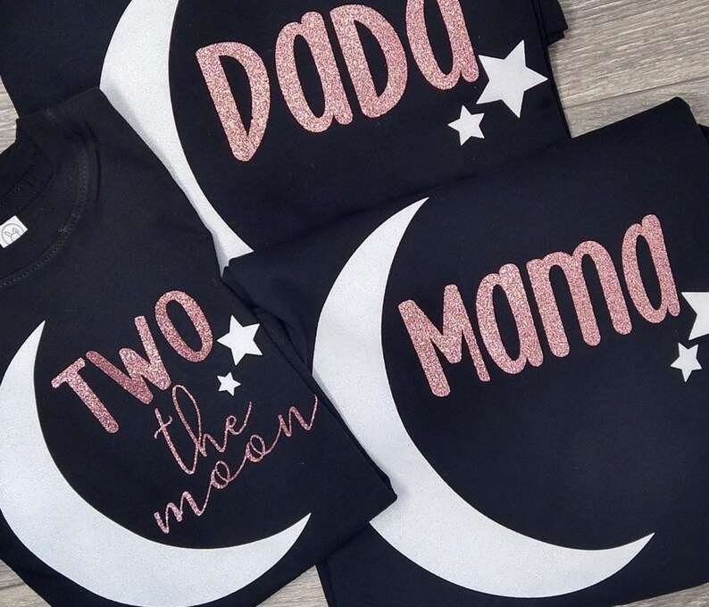 May include: Black t-shirts with white crescent moon and star designs. One shirt says "Two the moon" in pink glitter, another says "Dada", and a third says "Mama".
