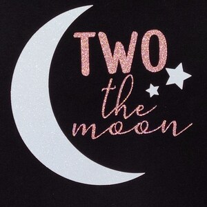 May include: Black t-shirt with a white glitter crescent moon and two glitter stars. The text "TWO the moon" is in pink glitter. The shirt is a cute design for a child's birthday.