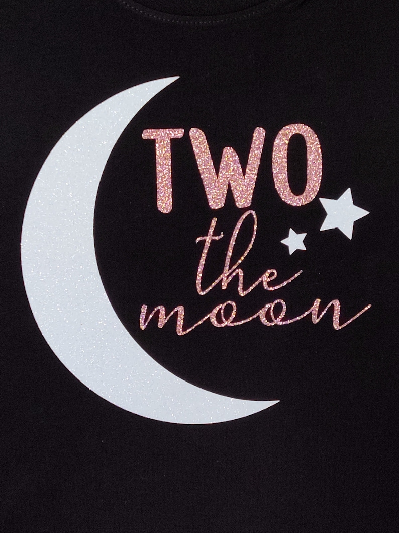 May include: Black t-shirt with a white glitter crescent moon and two glitter stars. The text "TWO the moon" is in pink glitter. The shirt is a cute design for a child's birthday.