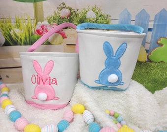 May include: Two Easter baskets with bunny designs. One basket is pink with the name Olivia embroidered on it. The other basket is blue. Both baskets are made of white fabric with a white fluffy tail on the bunny.