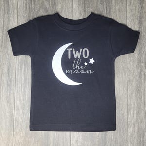 May include: Dark gray toddler t-shirt with a white crescent moon and the text "TWO the moon" in silver glitter. A small star is next to the text. The shirt is made of soft material.