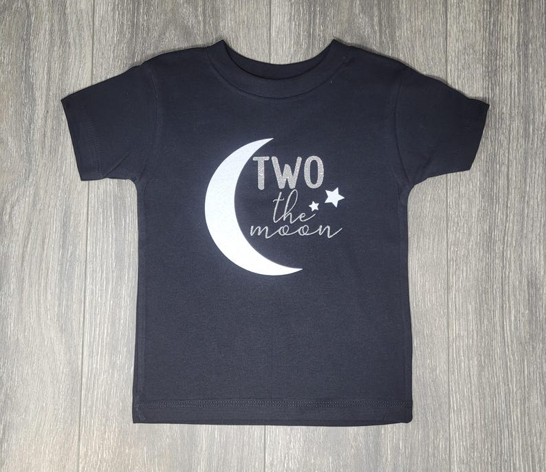 May include: Dark gray toddler t-shirt with a white crescent moon and the text "TWO the moon" in silver glitter. A small star is next to the text. The shirt is made of soft material.