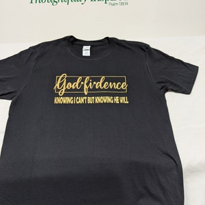May include: Black t-shirt with gold text that reads "God-fidence" in a rectangular box, with the text "Knowing I can't but knowing He will" below. The words "Thoughtfully Inspired" and "Psalm 139:14" are printed in green above the shirt.