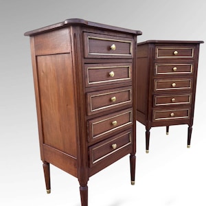 May include: Two antique wooden bedside tables. Each table has five drawers with gold-coloured trim and knobs. The tables are made of dark wood and have tapered legs with gold-coloured accents. The tables are set against a light grey background.