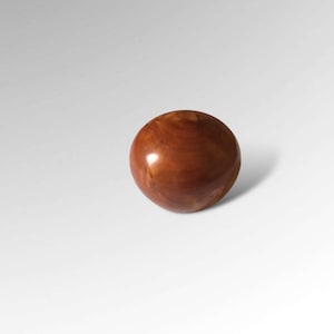 May include: A polished, spherical object with a wood-grain pattern. The ball is a warm brown colour and casts a shadow on the white surface. It appears to be a decorative item or a component of a larger piece, possibly furniture.