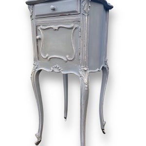May include: A grey painted bedside cabinet with decorative features. The small cabinet has a single drawer and a door, with carvings and curved legs. The finish has a distressed appearance, with silver accents. The cabinet is a classic piece of furniture, suitable for a bedroom.