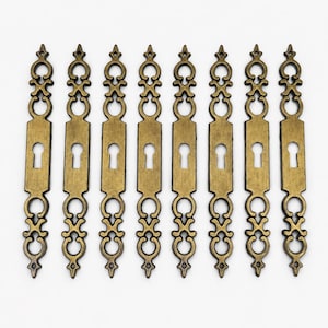 May include: Eight identical antique brass keyhole escutcheon plates arranged in a vertical row. Each metal plate features an elongated rectangular centre with a keyhole cutout, flanked by symmetrical, ornate scrollwork at the top and bottom ends.