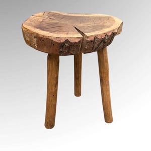 May include: A handcrafted wooden stool with three legs. The seat is a natural, irregular shape with a split, displaying the wood's grain and texture. The legs are simple, rounded, and attached to the seat, giving a rustic, sturdy appearance.