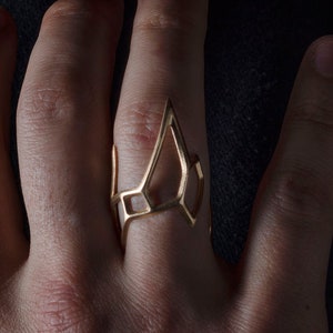 May include: A gold-toned geometric ring with a unique, open-work design. The ring is worn on the middle finger of a hand.