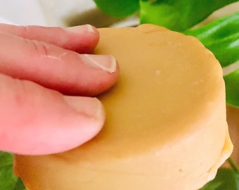 Può includere: Una saponetta rotonda di colore marrone chiaro. Il sapone è tenuto in una mano.