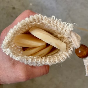 May include: A white cotton mesh bag with a wooden bead drawstring closure, containing multiple round, off-white soap bars.