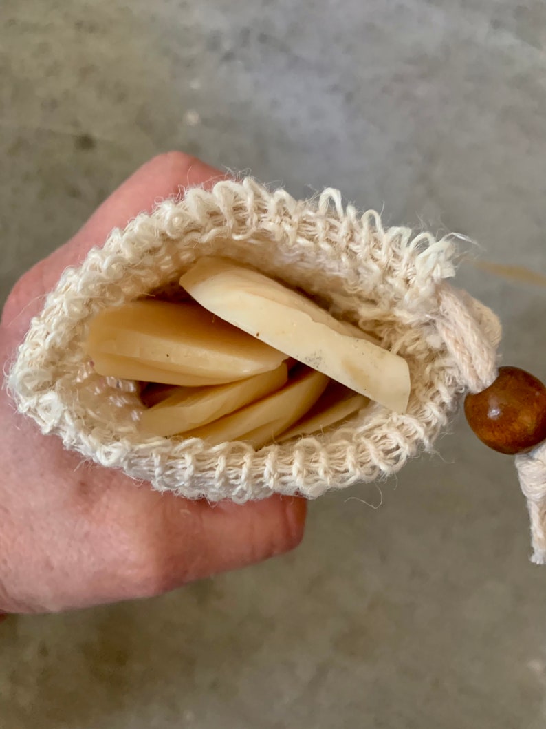 May include: A white cotton mesh bag with a wooden bead drawstring closure, containing multiple round, off-white soap bars.