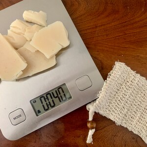 May include: A digital kitchen scale with a white bar of soap weighing 0.040 ounces. The soap is broken into several pieces. A white mesh soap bag is next to the scale.