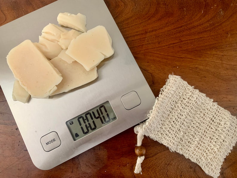 May include: A digital kitchen scale with a white bar of soap weighing 0.040 ounces. The soap is broken into several pieces. A white mesh soap bag is next to the scale.