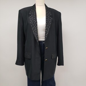 Könnte beinhalten: Schwarzer Blazer mit weißen Tupfen auf Revers und Taschen. Der Blazer hat goldfarbene Knöpfe und wird auf einer Schaufensterpuppe präsentiert. Er wird mit einem weißen Oberteil und dunkelblauen Jeans kombiniert.
