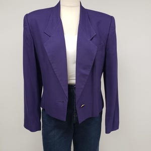 May include: A purple blazer with a subtle checkered pattern. The blazer has a notched lapel, a single-button closure, and a pocket on the left side. The blazer is paired with blue jeans and a white top.
