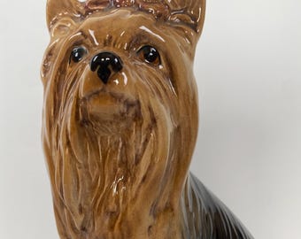 Könnte beinhalten: Eine Keramikfigur eines Yorkshire Terriers in sitzender Position. Der Hund hat braunes und schwarzes Fell mit glänzendem Finish. Die Figur hat detaillierte Merkmale, einschließlich einer kleinen Schleife auf dem Kopf und einem aufmerksamen Ausdruck.