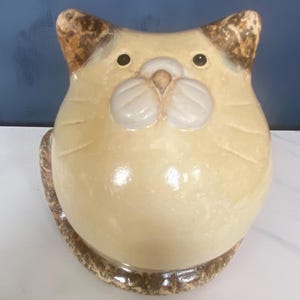 May include: A ceramic cat ornament with a rounded body and a light yellow glaze. The cat has brown-tipped ears, black eyes, and a white muzzle. The base is a textured brown. A decorative item.