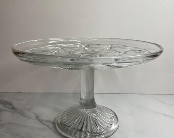 Könnte beinhalten: Ein klarer Kuchenständer aus Glas mit einer runden, leicht gewellten Oberseite. Die Oberseite hat ein Blumenmuster. Der Ständer hat einen Sockel mit vertikalen Rippen. Der Stiel ist glatt und zylindrisch. Der Ständer steht auf einer weißen Marmoroberfläche.