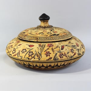 May include: A round ceramic lidded bowl with a cream-colored base featuring black and red hand-painted floral and bird motifs. The lid has a black knob handle and a Greek key border pattern, while the base displays a repeating black triangular design.