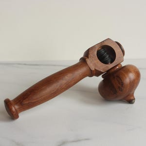 May include: A brown wooden nutcracker tool featuring a long, smooth handle and a rounded, bulbous head with a circular opening that contains a dark, threaded metal screw mechanism for cracking nuts with a simple twisting motion.