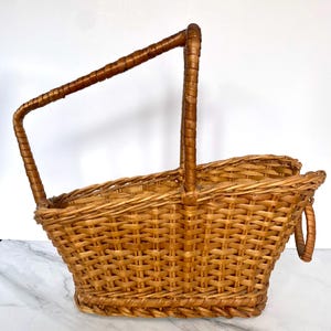 K&ouml;nnte beinhalten: Ein hellbrauner Picknickkorb aus geflochtenem Korbgeflecht mit einem hohen, gebogenen Griff und einem kleineren Griff an der Seite. Der Korb hat eine rechteckige Form mit einer leicht ausgestellten Oberseite. Der Griff ist mit dunklerem braunem Material umwickelt.
