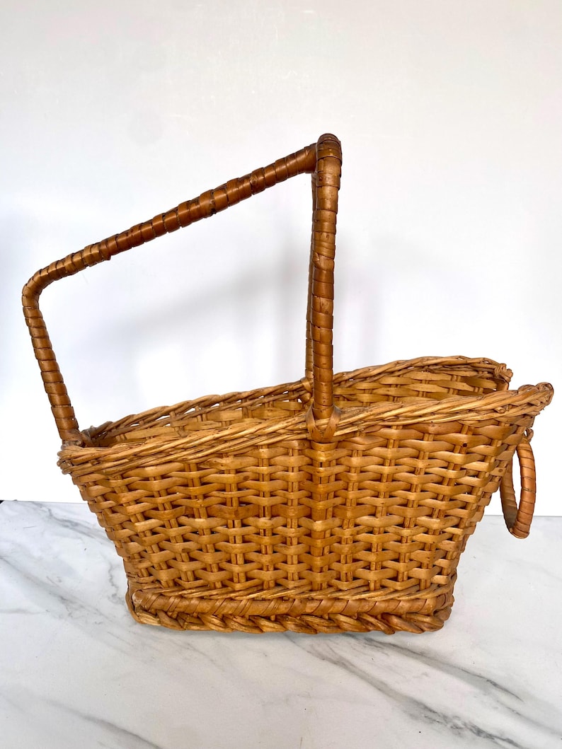 K&ouml;nnte beinhalten: Ein hellbrauner Picknickkorb aus geflochtenem Korbgeflecht mit einem hohen, gebogenen Griff und einem kleineren Griff an der Seite. Der Korb hat eine rechteckige Form mit einer leicht ausgestellten Oberseite. Der Griff ist mit dunklerem braunem Material umwickelt.
