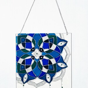 Op de afbeelding: Een rechthoekig glas-in-loodpaneel met een geometrisch ontwerp in blauwe, groene en zilveren tinten. Opgeschort aan een zilveren ketting. Versierd met hangende groene en transparante kralen.