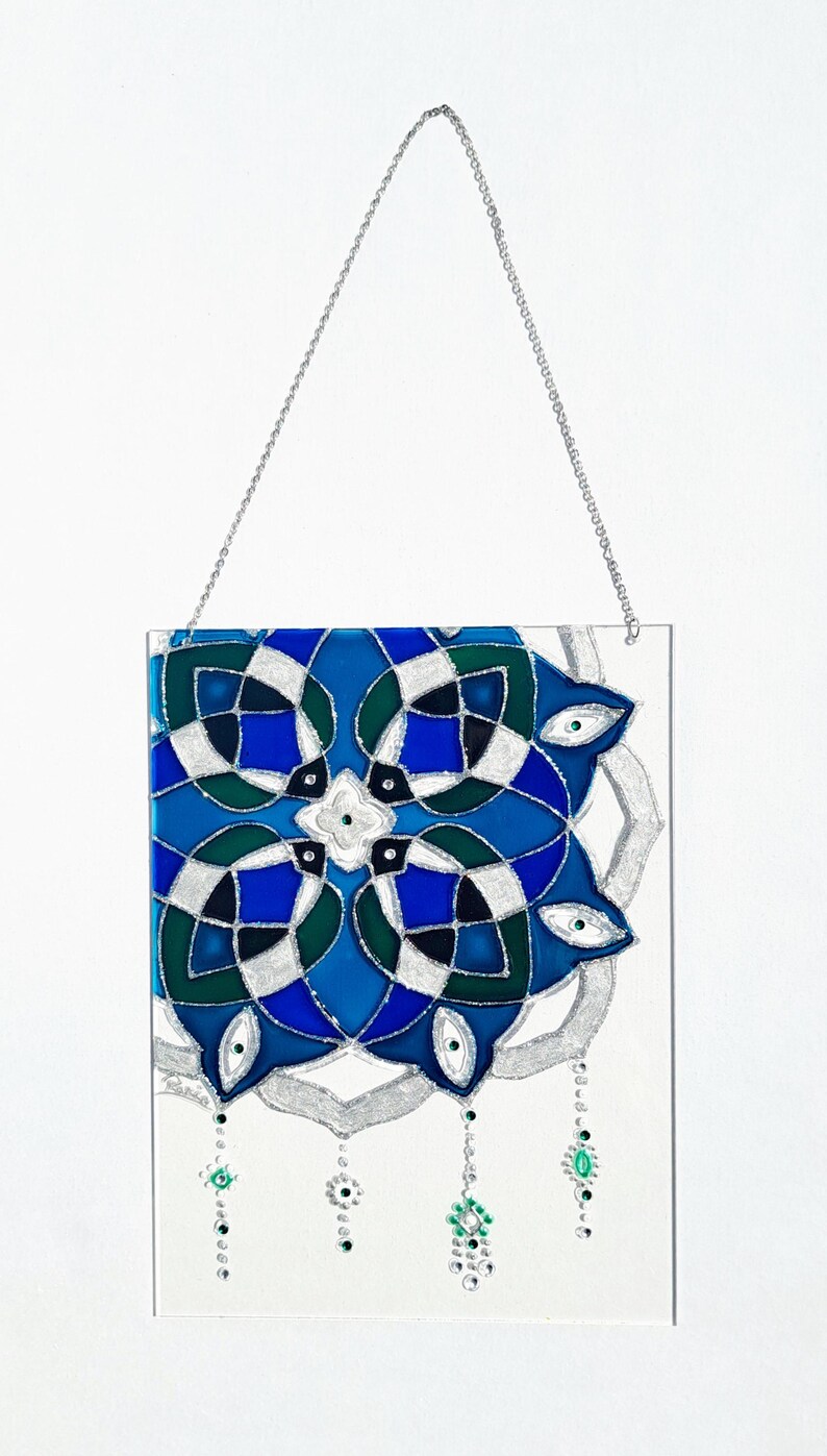 Op de afbeelding: Een rechthoekig glas-in-loodpaneel met een geometrisch ontwerp in blauwe, groene en zilveren tinten. Opgeschort aan een zilveren ketting. Versierd met hangende groene en transparante kralen.