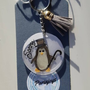 Op de afbeelding: Een witte en grijze pingu&iuml;n met een zwarte hoge hoed en een zwarte stok. De pingu&iuml;n zit op een ronde, doorzichtige acryl sleutelhanger met de naam "Sonny" erop. De sleutelhanger is bevestigd aan een zilveren ketting en een grijze kwast. De sleutelhanger heeft ook een witte sticker met de tekst "Craft Shop @ Eot" en "Pariajoys" erop.