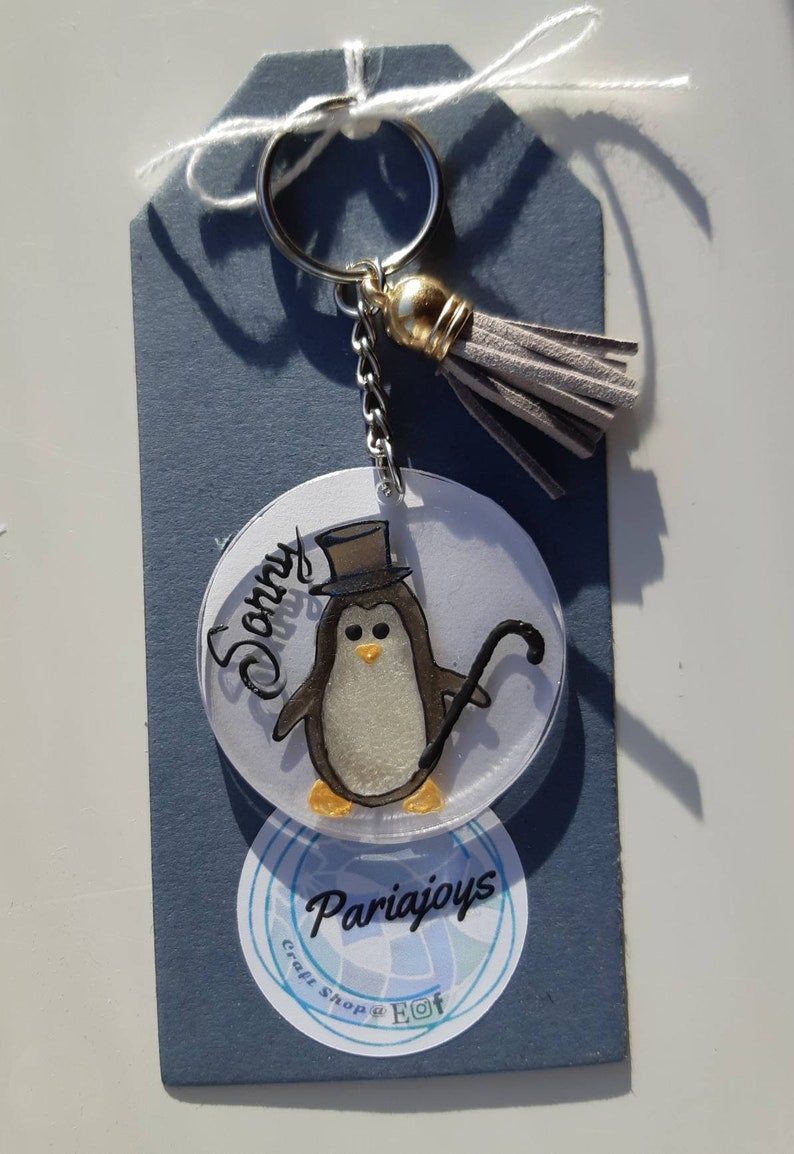 Op de afbeelding: Een witte en grijze pingu&iuml;n met een zwarte hoge hoed en een zwarte stok. De pingu&iuml;n zit op een ronde, doorzichtige acryl sleutelhanger met de naam "Sonny" erop. De sleutelhanger is bevestigd aan een zilveren ketting en een grijze kwast. De sleutelhanger heeft ook een witte sticker met de tekst "Craft Shop @ Eot" en "Pariajoys" erop.