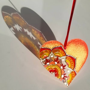 Op de afbeelding: Een rode hartvormige ornament met een gouden en oranje mandala-ontwerp. De ornament hangt aan een rood touwtje.