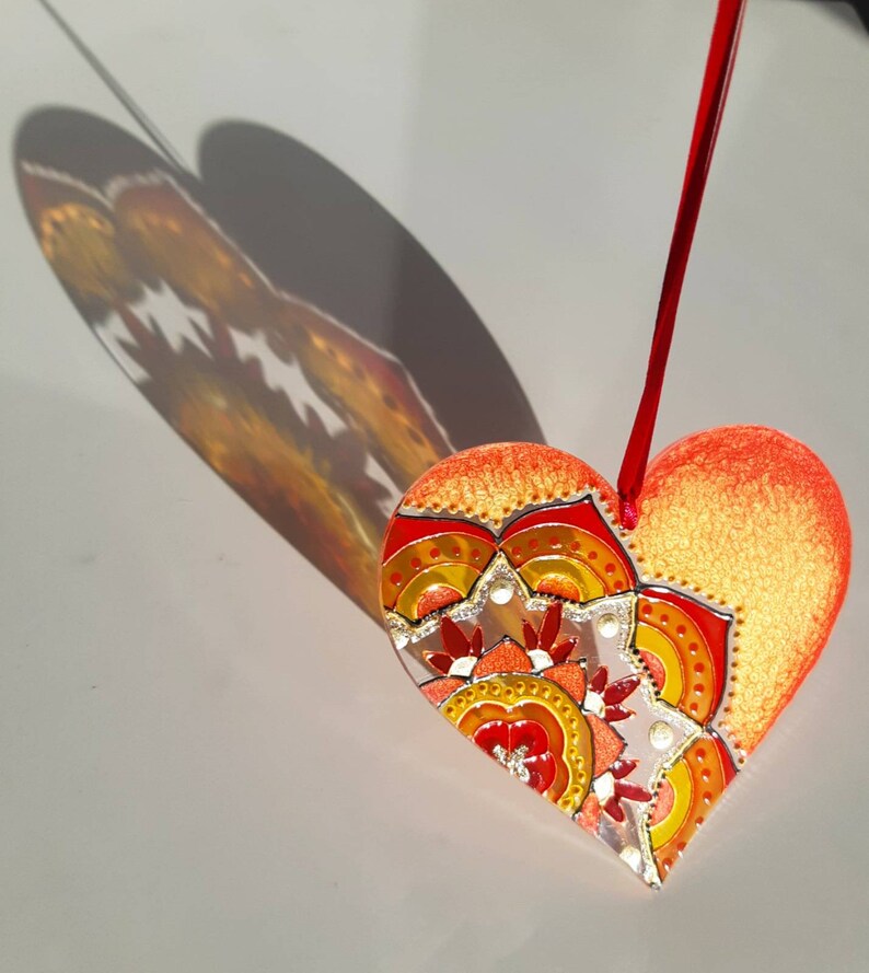 Op de afbeelding: Een rode hartvormige ornament met een gouden en oranje mandala-ontwerp. De ornament hangt aan een rood touwtje.