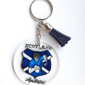 Op de afbeelding: Een ronde, transparante acryl sleutelhanger met een blauw en wit Schots vlagontwerp en een rugby-speler die een bal vasthoudt. De sleutelhanger heeft een zilveren ring en een blauwe kwast. De tekst "Scotland" en "Andrew" staat op de sleutelhanger.