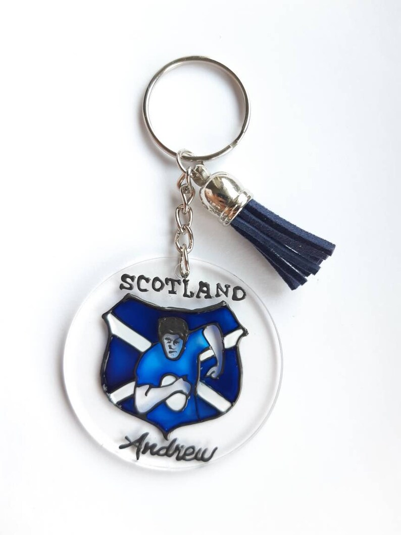 Op de afbeelding: Een ronde, transparante acryl sleutelhanger met een blauw en wit Schots vlagontwerp en een rugby-speler die een bal vasthoudt. De sleutelhanger heeft een zilveren ring en een blauwe kwast. De tekst "Scotland" en "Andrew" staat op de sleutelhanger.