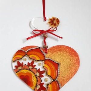 Op de afbeelding: Een hartvormige hanger in rood en oranje met een mandala-ontwerp. De hanger is gemaakt van glas en heeft een gouden glitterafwerking. Hij hangt aan een rood lint.