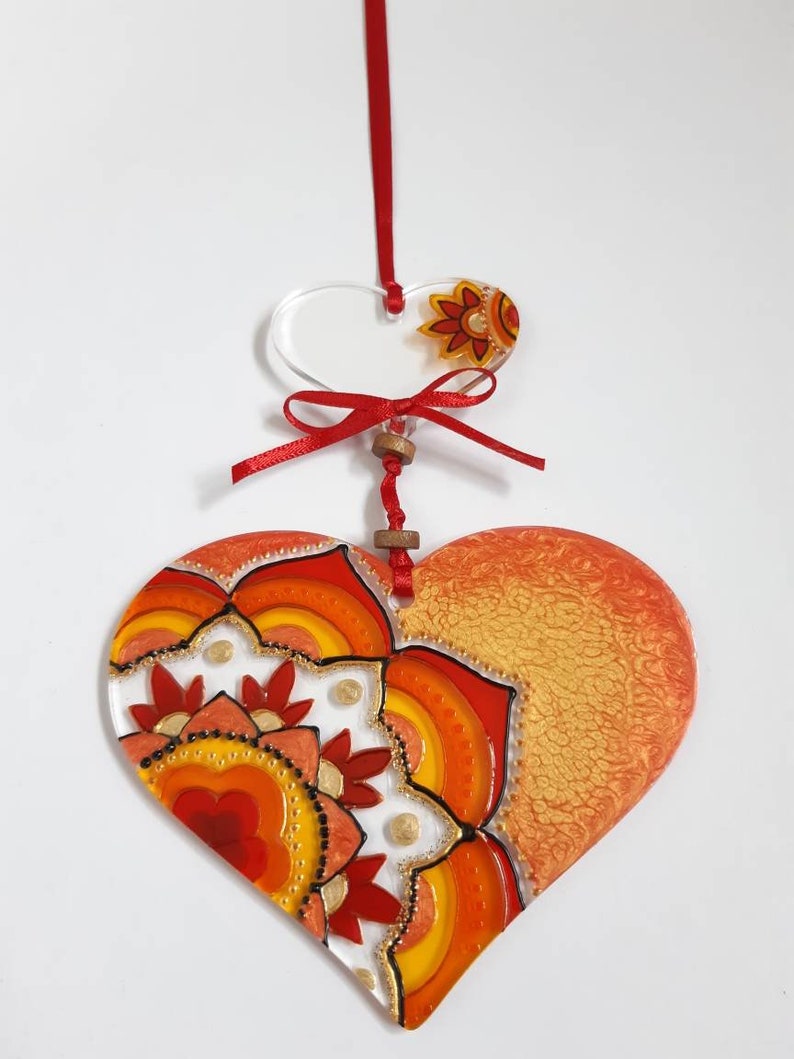 Op de afbeelding: Een hartvormige hanger in rood en oranje met een mandala-ontwerp. De hanger is gemaakt van glas en heeft een gouden glitterafwerking. Hij hangt aan een rood lint.