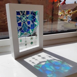 Op de afbeelding: Een ingelijst glas-in-lood kunstwerk met een blauw en groen bloemmotief. Het kunstwerk wordt gepresenteerd in een witte lijst en werpt een schaduw op een wit oppervlak. Het stuk wordt verlicht door zonlicht.