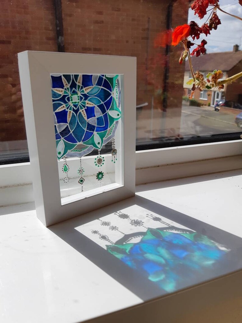 Op de afbeelding: Een ingelijst glas-in-lood kunstwerk met een blauw en groen bloemmotief. Het kunstwerk wordt gepresenteerd in een witte lijst en werpt een schaduw op een wit oppervlak. Het stuk wordt verlicht door zonlicht.