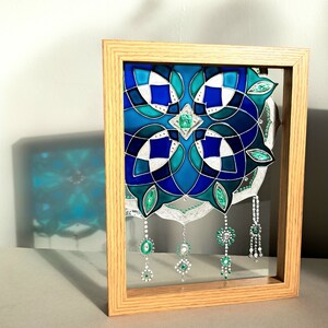 Op de afbeelding: Een ingelijst glas-in-lood kunstwerk met een geometrisch ontwerp in tinten blauw, turquoise en wit. Het kunstwerk bevat hangende juweelachtige versieringen in groen en zilver, geplaatst in een natuurlijke houten lijst.